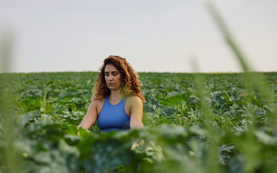 selective focus photography of woman meditating while sitting on ground surrounded by plants during daytime